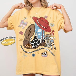 May include: A mustard yellow t-shirt featuring a graphic print of a cowboy boot with a cow print pattern, a red hat, a female character, and blocks with the letters T, S, and B. The text Taylor and I KNEW IT I KNEW YOU are printed on the shirt.