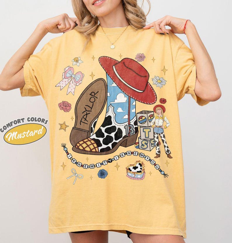 May include: A mustard yellow t-shirt featuring a graphic print of a cowboy boot with a cow print pattern, a red hat, a female character, and blocks with the letters T, S, and B. The text Taylor and I KNEW IT I KNEW YOU are printed on the shirt.