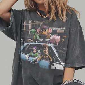 Puede incluir: Una camiseta gris carb&oacute;n de corte oversize con un estampado gr&aacute;fico de personajes animados, incluyendo una patata, un dinosaurio verde, un cerdo rosa, un ranger espacial y un vaquero asomados a un coche azul claro. El texto dice TOY SORANCSTHQS.