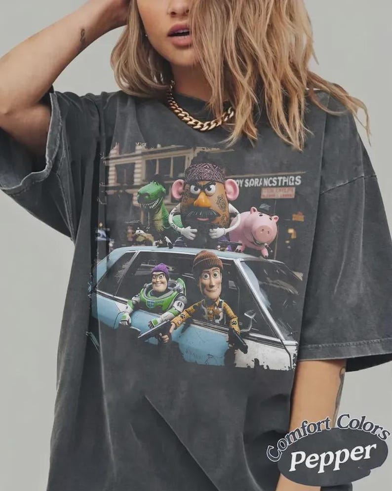 Puede incluir: Una camiseta gris carb&oacute;n de corte oversize con un estampado gr&aacute;fico de personajes animados, incluyendo una patata, un dinosaurio verde, un cerdo rosa, un ranger espacial y un vaquero asomados a un coche azul claro. El texto dice TOY SORANCSTHQS.