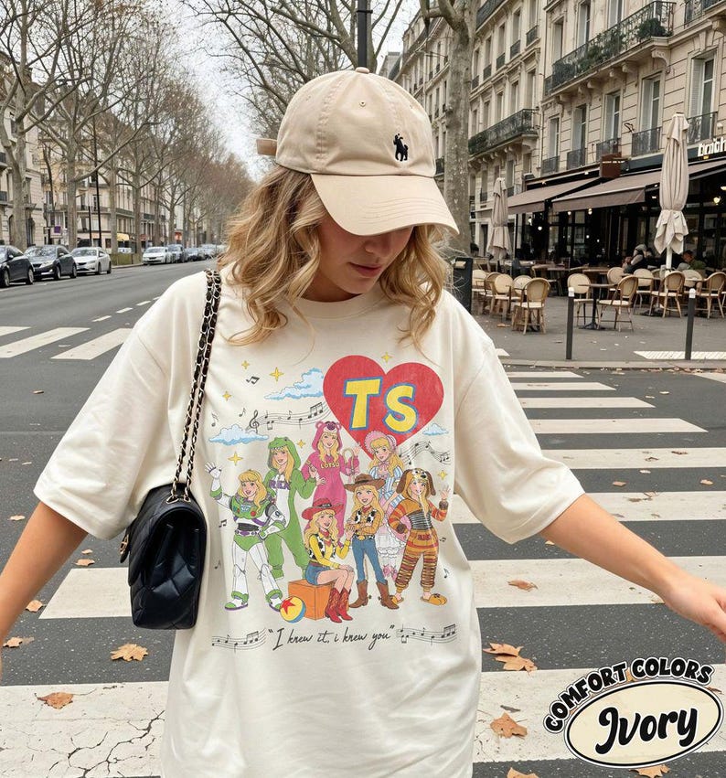 Puede incluir: Una camiseta color marfil con un estampado gr&aacute;fico de varios personajes disfrazados, un coraz&oacute;n rojo con las letras TS y el texto "I knew it, I knew you" con notas musicales. La camiseta se combina con una gorra de b&eacute;isbol beige y un bolso negro.