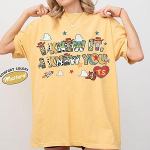 May include: A mustard yellow short-sleeve t-shirt featuring a graphic print with the text I KNEW IT, I KNEW YOU. The design includes cowboy hats, rockets, clouds, stars, and a red heart with the letters TS, incorporating patterns from toy characters.