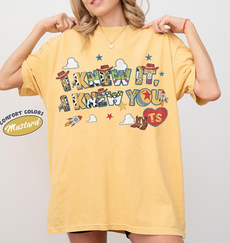 May include: A mustard yellow short-sleeve t-shirt featuring a graphic print with the text I KNEW IT, I KNEW YOU. The design includes cowboy hats, rockets, clouds, stars, and a red heart with the letters TS, incorporating patterns from toy characters.