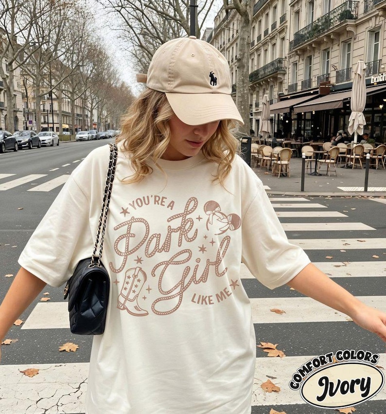 Op de afbeelding: Een ivoorkleurig T-shirt met korte mouwen en een bruine grafische print met de tekst "You're a Park Girl Like Me", een cowboylaars en muizenoren. Het shirt wordt gecombineerd met een beige honkbalpet met een klein donker logo en een zwarte schoudertas.