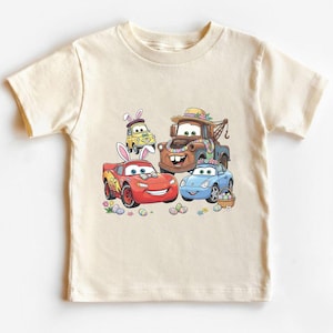 Può includere: Una maglietta color crema con una grafica colorata di auto animate decorate per Pasqua. Le auto includono un'auto da corsa rossa, un'auto sportiva blu, un'auto gialla e un carro attrezzi marrone, tutte con accessori pasquali.