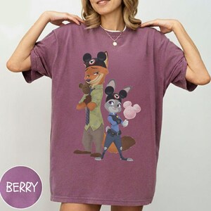 Pu&ograve; includere: T-shirt color bacca con un'illustrazione di Nick Wilde e Judy Hopps di Zootropolis, entrambi con le orecchie di Topolino. Nick tiene un orsacchiotto e Judy tiene lo zucchero filato. La parola "BERRY" &egrave; in un cerchio in basso.