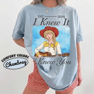 May include: A light blue t-shirt featuring a graphic of a blonde individual in a red cowboy hat playing an acoustic guitar. The text reads EST 2026 I Knew It I Knew You with a treble clef symbol. A Comfort Colors Chambray label is visible.