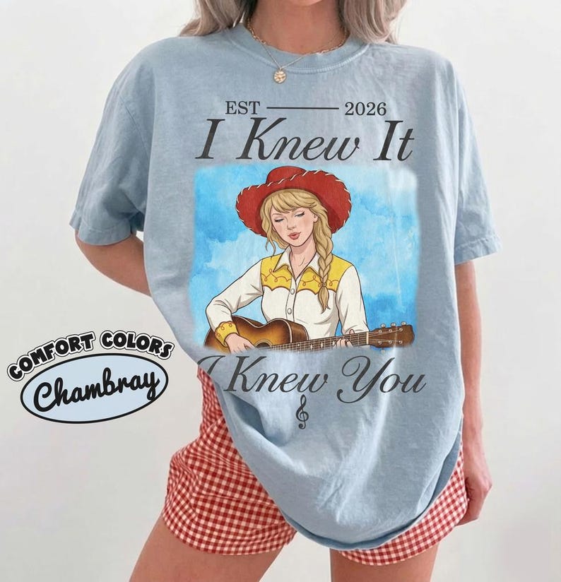 May include: A light blue t-shirt featuring a graphic of a blonde individual in a red cowboy hat playing an acoustic guitar. The text reads EST 2026 I Knew It I Knew You with a treble clef symbol. A Comfort Colors Chambray label is visible.