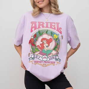 May include: A lilac t-shirt with a graphic of Ariel from The Little Mermaid. The design shows Ariel in a seashell, with the word "ARIEL" above and "EST. 1989 DISNEY PRINCESS" below. The shirt has short sleeves.