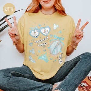 May include: A yellow t-shirt featuring a Disney Elsa design. The design includes images of Elsa, Olaf, and other Frozen-themed elements. The text "Disney Elsa" is also present on the shirt.