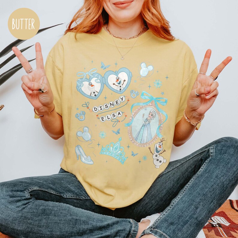 May include: A yellow t-shirt featuring a Disney Elsa design. The design includes images of Elsa, Olaf, and other Frozen-themed elements. The text "Disney Elsa" is also present on the shirt.
