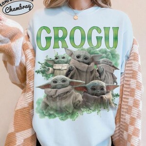May include: A light blue crewneck sweatshirt featuring a graphic print of multiple Grogu characters in beige robes surrounded by green smoke. The text GROGU is printed in large green letters above the characters. A Comfort Colors Chambray logo is visible.