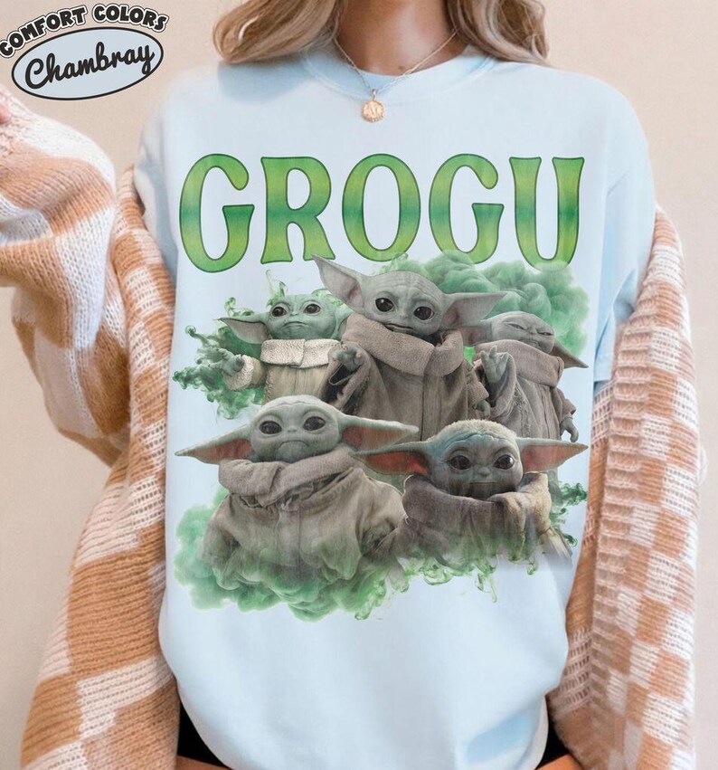 May include: A light blue crewneck sweatshirt featuring a graphic print of multiple Grogu characters in beige robes surrounded by green smoke. The text GROGU is printed in large green letters above the characters. A Comfort Colors Chambray logo is visible.