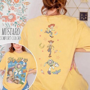 May include: Mustard-colored t-shirt with a Toy Story graphic on the front and back. The front features the Toy Story logo and characters. The back has illustrations of Jessie, Woody, and Buzz Lightyear. The shirt is labeled "Comfort Colors."