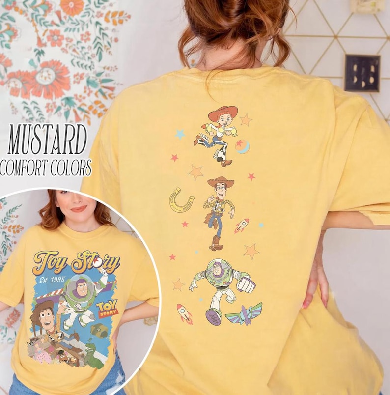 May include: Mustard-colored t-shirt with a Toy Story graphic on the front and back. The front features the Toy Story logo and characters. The back has illustrations of Jessie, Woody, and Buzz Lightyear. The shirt is labeled "Comfort Colors."
