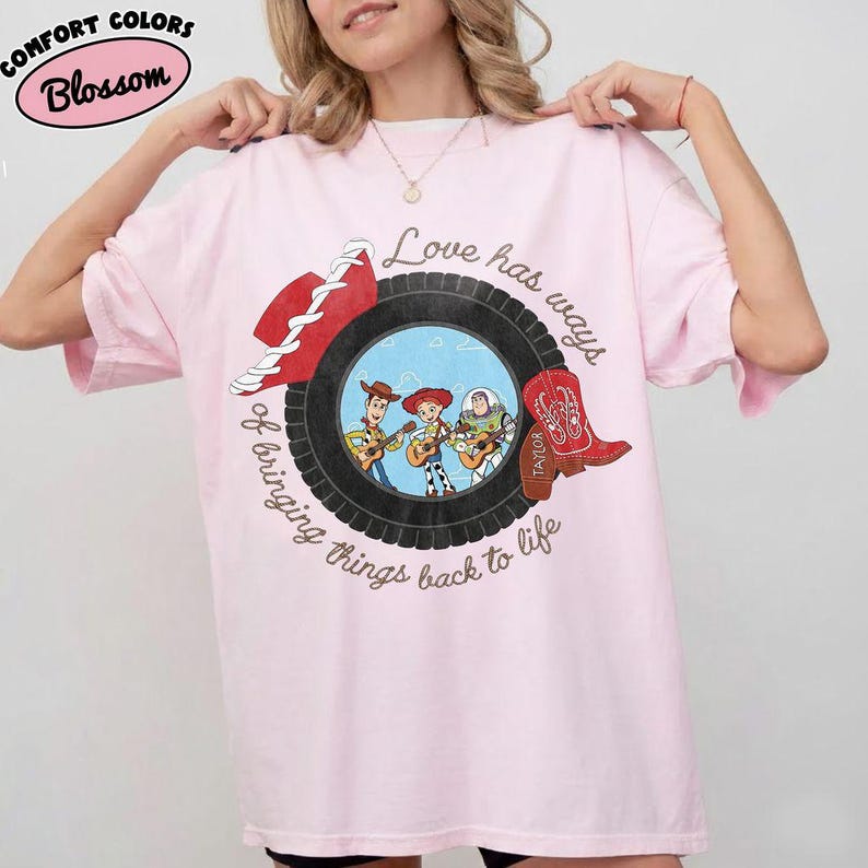 May include: A light pink t-shirt featuring a graphic of a black tire framing three cartoon characters playing guitars. A red cowboy hat and a red cowboy boot are positioned on the tire. The text reads, Love has ways of bringing things back to life.