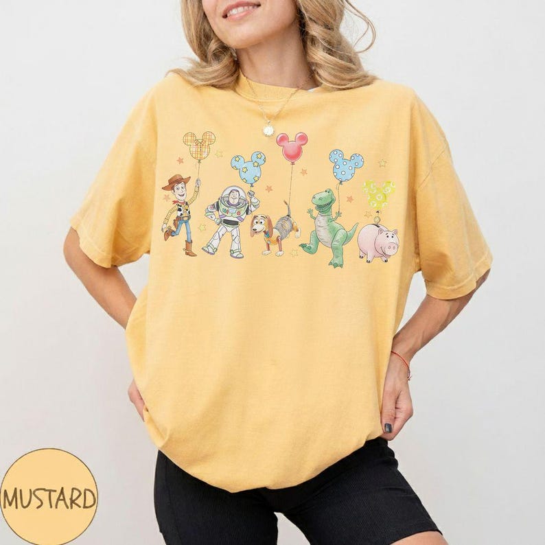 May include: Mustard-colored t-shirt featuring a graphic of cartoon characters holding Mickey Mouse-shaped balloons. The characters include a cowboy, a space ranger, a dog, a dinosaur, and a pig. The shirt has a crew neck and short sleeves.
