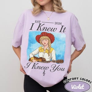 May include: A light purple t-shirt featuring a graphic of a person with blonde hair wearing a red cowboy hat and a yellow and white shirt while playing an acoustic guitar. The text reads EST 2026 I Knew It I Knew You with a treble clef symbol.