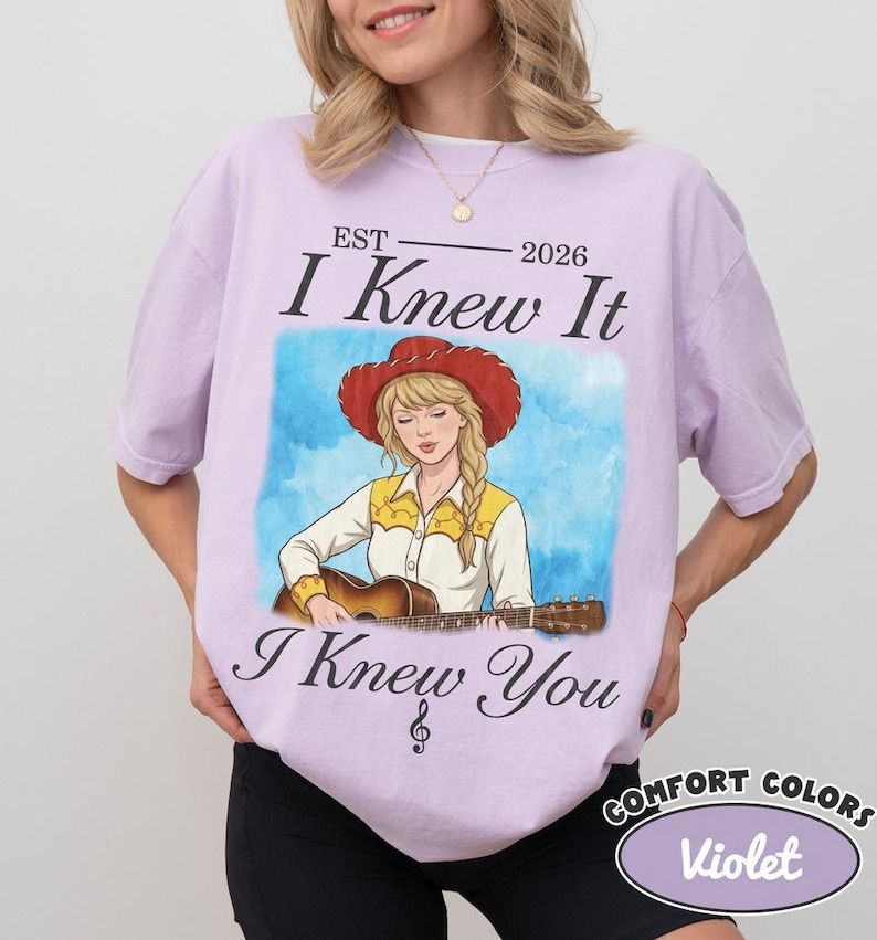 May include: A light purple t-shirt featuring a graphic of a person with blonde hair wearing a red cowboy hat and a yellow and white shirt while playing an acoustic guitar. The text reads EST 2026 I Knew It I Knew You with a treble clef symbol.