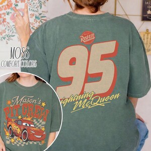 Op de afbeelding: Een mosgroen t-shirt met een grote afbeelding op de achterkant met het nummer 95 in beige met een rode omlijning, het Rust-eze logo en de tekst Lightning McQueen. De voorkant toont een rode raceauto met de tekst Mason's Pit Crew.