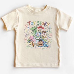 K&ouml;nnte beinhalten: Cremefarbenes T-Shirt mit einer farbenfrohen Grafik von Figuren aus dem Animationsfilm "Toy Story". Das Design enth&auml;lt die Worte "Toy Story" in einer verspielten Schriftart sowie Zeichnungen von Woody, Buzz Lightyear und anderen Charakteren.
