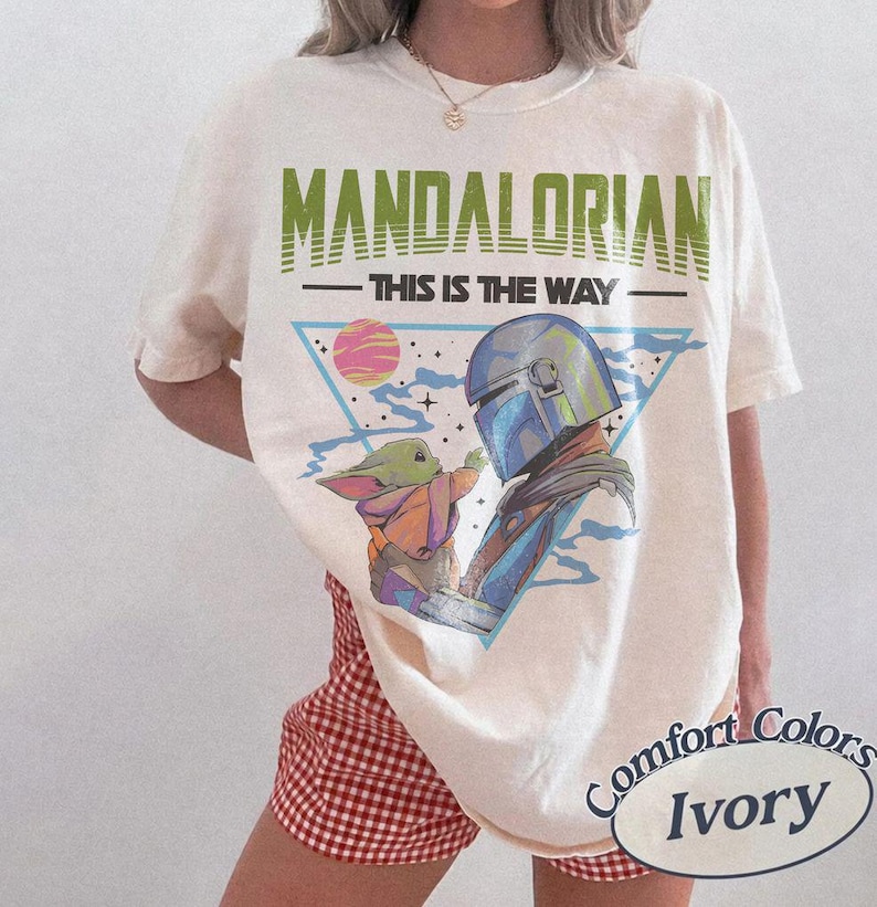 Może przedstawiać: T-shirt w kolorze kości słoniowej z nadrukiem graficznym przedstawiającym Mandalorianina i Grogu. Tekst głosi MANDALORIAN oraz THIS IS THE WAY. Wz&oacute;r zawiera niebieski tr&oacute;jkąt, r&oacute;żową planetę i gwiazdy. Koszulka ma okrągły dekolt i kr&oacute;tkie rękawy.