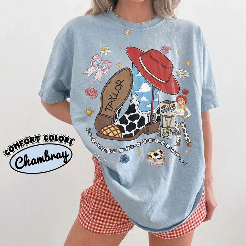 May include: A light blue t-shirt featuring a graphic print of a cowboy boot with a cow print pattern, a red hat, a character, and blocks with the letters T, S, and S. The text TAYLOR is on the boot, and the text I KNEW IT I KNEW YOU is on a string.