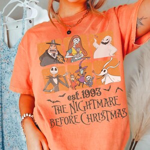 May include: Orange t-shirt with characters from "The Nightmare Before Christmas." The design includes Jack Skellington, Sally, and other characters, with the text "est. 1993" and "The Nightmare Before Christmas."