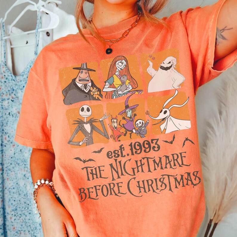 May include: Orange t-shirt with characters from "The Nightmare Before Christmas." The design includes Jack Skellington, Sally, and other characters, with the text "est. 1993" and "The Nightmare Before Christmas."