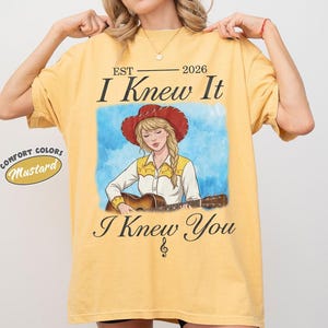 May include: A mustard yellow t-shirt featuring a graphic of a blonde person in a red cowboy hat playing an acoustic guitar. The text reads EST 2026 I Knew It I Knew You with a treble clef symbol. A small circular label reads COMFORT COLORS Mustard.
