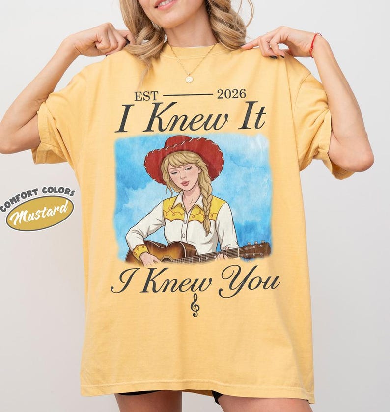 May include: A mustard yellow t-shirt featuring a graphic of a blonde person in a red cowboy hat playing an acoustic guitar. The text reads EST 2026 I Knew It I Knew You with a treble clef symbol. A small circular label reads COMFORT COLORS Mustard.