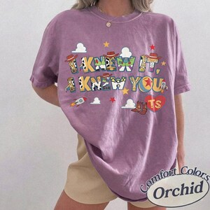 May include: A short-sleeve orchid purple t-shirt featuring a graphic print with the text I KNEW IT, I KNEW YOU. The design includes colorful letters patterned with cowboy and space ranger motifs, small clouds, stars, a rocket, and a red heart with TS.