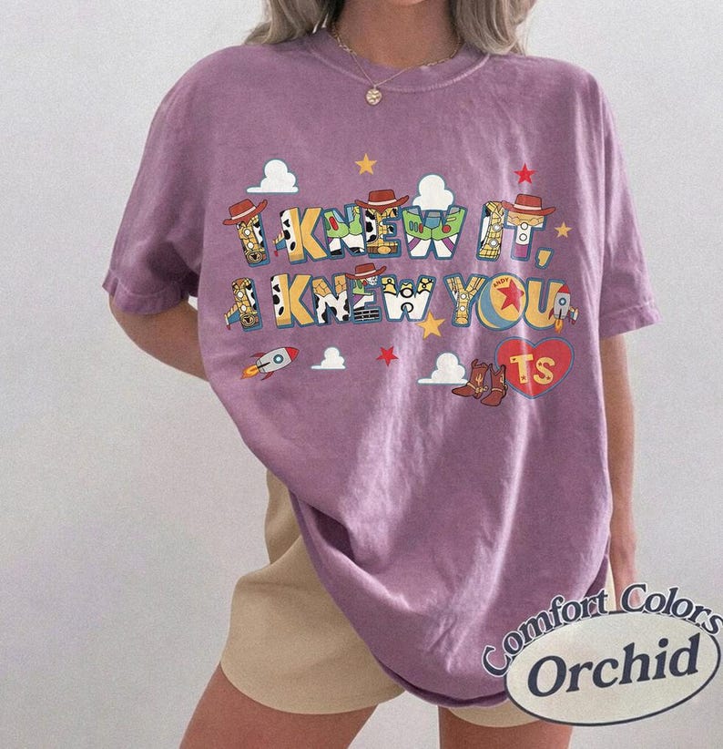 May include: A short-sleeve orchid purple t-shirt featuring a graphic print with the text I KNEW IT, I KNEW YOU. The design includes colorful letters patterned with cowboy and space ranger motifs, small clouds, stars, a rocket, and a red heart with TS.