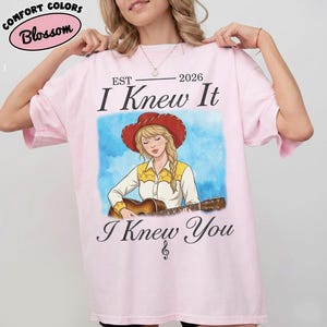 May include: A light pink t-shirt featuring a graphic of a person in a red cowboy hat playing an acoustic guitar. The text reads EST 2026, I Knew It, and I Knew You with a treble clef symbol. The Comfort Colors Blossom logo is visible in the corner.