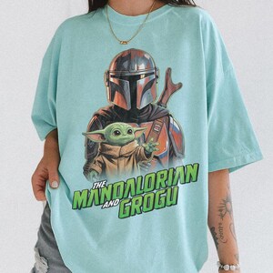 May include: A light mint green t-shirt featuring a graphic print of The Mandalorian and Grogu. The design displays the armored character behind the small green creature, with the text "THE MANDALORIAN AND GROGU" printed in bold green letters below.