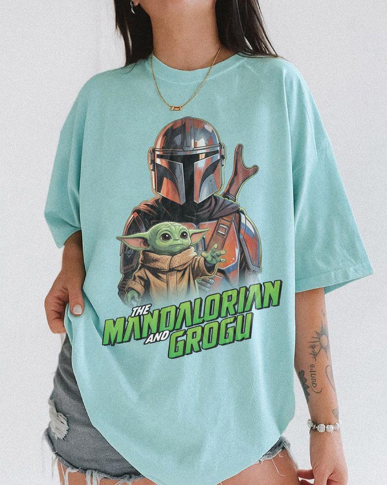 May include: A light mint green t-shirt featuring a graphic print of The Mandalorian and Grogu. The design displays the armored character behind the small green creature, with the text "THE MANDALORIAN AND GROGU" printed in bold green letters below.