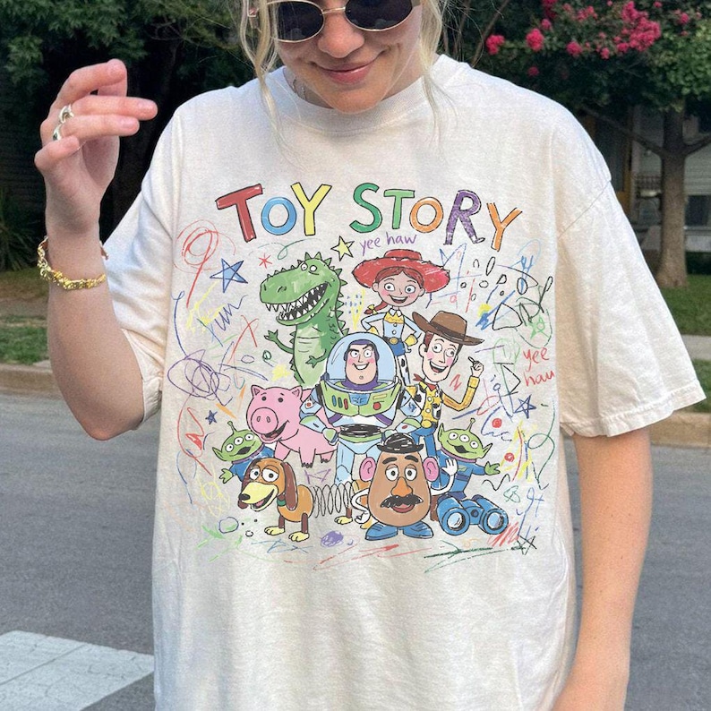 K&ouml;nnte beinhalten: Cremefarbenes T-Shirt mit dem Schriftzug "TOY STORY" in bunten Buchstaben &uuml;ber einer Grafik von Figuren aus dem Animationsfilm. Das Design umfasst Woody, Buzz Lightyear und andere Charaktere mit bunten Kritzeleien.