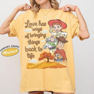 May include: A mustard yellow short-sleeve t-shirt featuring a graphic print of Jessie, Buzz Lightyear, and Woody from Toy Story. The text reads, Love has ways of bringing things back to life, above an illustration of a tree on a hill.