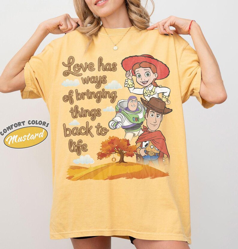 May include: A mustard yellow short-sleeve t-shirt featuring a graphic print of Jessie, Buzz Lightyear, and Woody from Toy Story. The text reads, Love has ways of bringing things back to life, above an illustration of a tree on a hill.