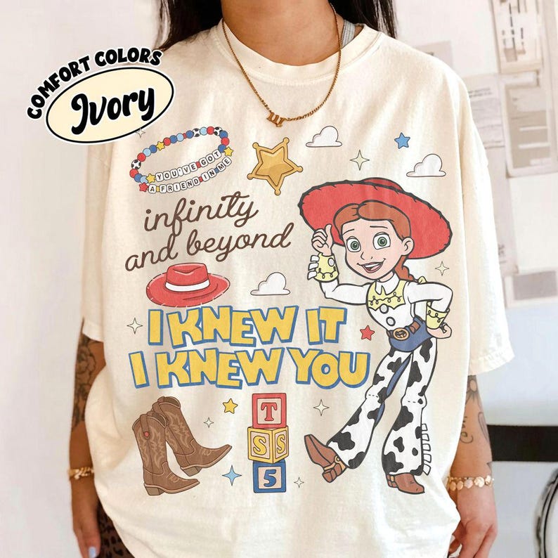 May include: An ivory t-shirt featuring a graphic print of a cowgirl character, a red cowboy hat, brown cowboy boots, a beaded bracelet, a star, and alphabet blocks. The text reads: You've got a friend in me, infinity and beyond, I knew it, I knew you.