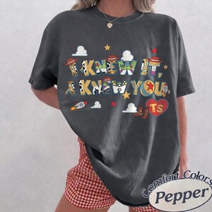 May include: A charcoal gray short-sleeve t-shirt featuring a graphic print with the text I KNEW IT, I KNEW YOU. The letters are filled with patterns inspired by toy characters, including cow print, space ranger colors, and a ball. Includes clouds and stars.