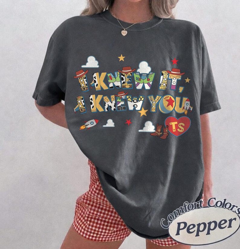 May include: A charcoal gray short-sleeve t-shirt featuring a graphic print with the text I KNEW IT, I KNEW YOU. The letters are filled with patterns inspired by toy characters, including cow print, space ranger colors, and a ball. Includes clouds and stars.