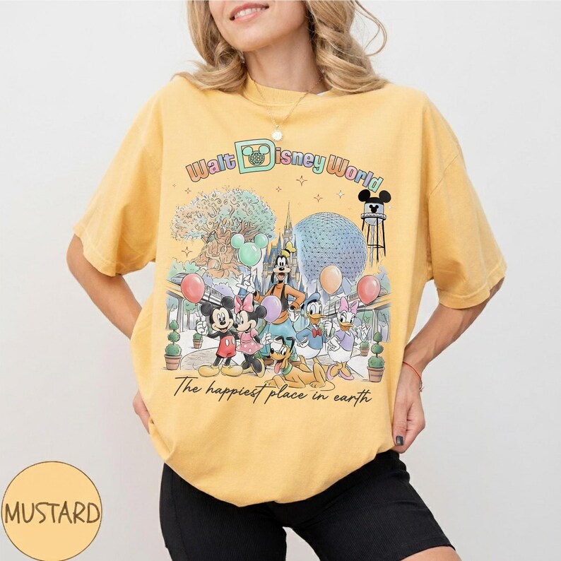 May include: Mustard yellow t-shirt featuring a colorful graphic of Disney characters at Walt Disney World. The design includes Mickey and Minnie Mouse, Goofy, Donald and Daisy Duck, and the text "The happiest place on earth."