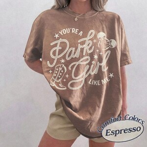 Op de afbeelding: Een bruin t-shirt met korte mouwen en een cr&egrave;mekleurige grafische print met de tekst "YOU'RE A Park Girl LIKE ME". Het ontwerp bevat een cowboylaars, een strik en sterren. Het t-shirt heeft een ronde hals en een relaxte pasvorm.