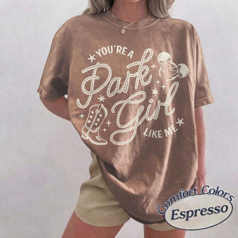 Op de afbeelding: Een bruin t-shirt met korte mouwen en een cr&egrave;mekleurige grafische print met de tekst "YOU'RE A Park Girl LIKE ME". Het ontwerp bevat een cowboylaars, een strik en sterren. Het t-shirt heeft een ronde hals en een relaxte pasvorm.