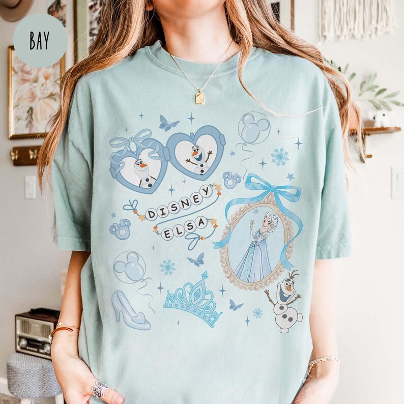 May include: Light blue t-shirt featuring Disney's Elsa. The shirt has a graphic design with sunglasses, snowflakes, and the word "Disney Elsa". The design includes images of Olaf, a crown, and a high-heeled shoe.