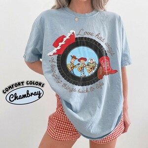 May include: A light blue t-shirt featuring a graphic of a black tire surrounding an illustration of three animated characters playing guitars. The design includes a red cowboy hat, a red cowboy boot with the name Taylor, and the text Love has ways of bringing things back to life.