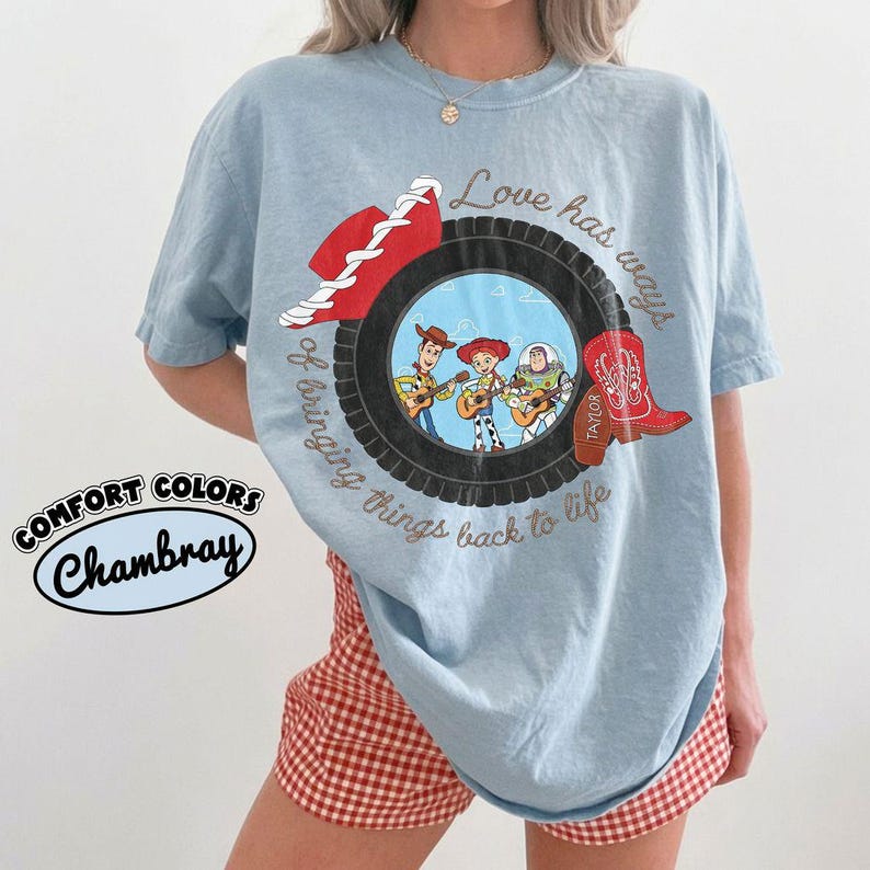 May include: A light blue t-shirt featuring a graphic of a black tire surrounding an illustration of three animated characters playing guitars. The design includes a red cowboy hat, a red cowboy boot with the name Taylor, and the text Love has ways of bringing things back to life.