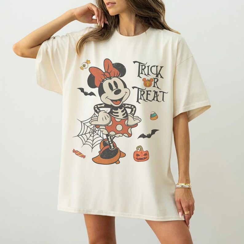 May include: An off-white short-sleeve t-shirt featuring a graphic of a skeleton Minnie Mouse wearing a polka dot skirt and bow. The design includes the text "Trick or Treat" alongside illustrations of bats, a spider web, a pumpkin, and candy.