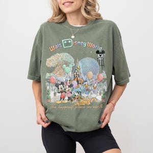 May include: Olive green t-shirt featuring a colorful graphic of Disney characters and landmarks, including Mickey Mouse, Minnie Mouse, and the Spaceship Earth. The text "Walt Disney World" and "The happiest place in earth" are also displayed.